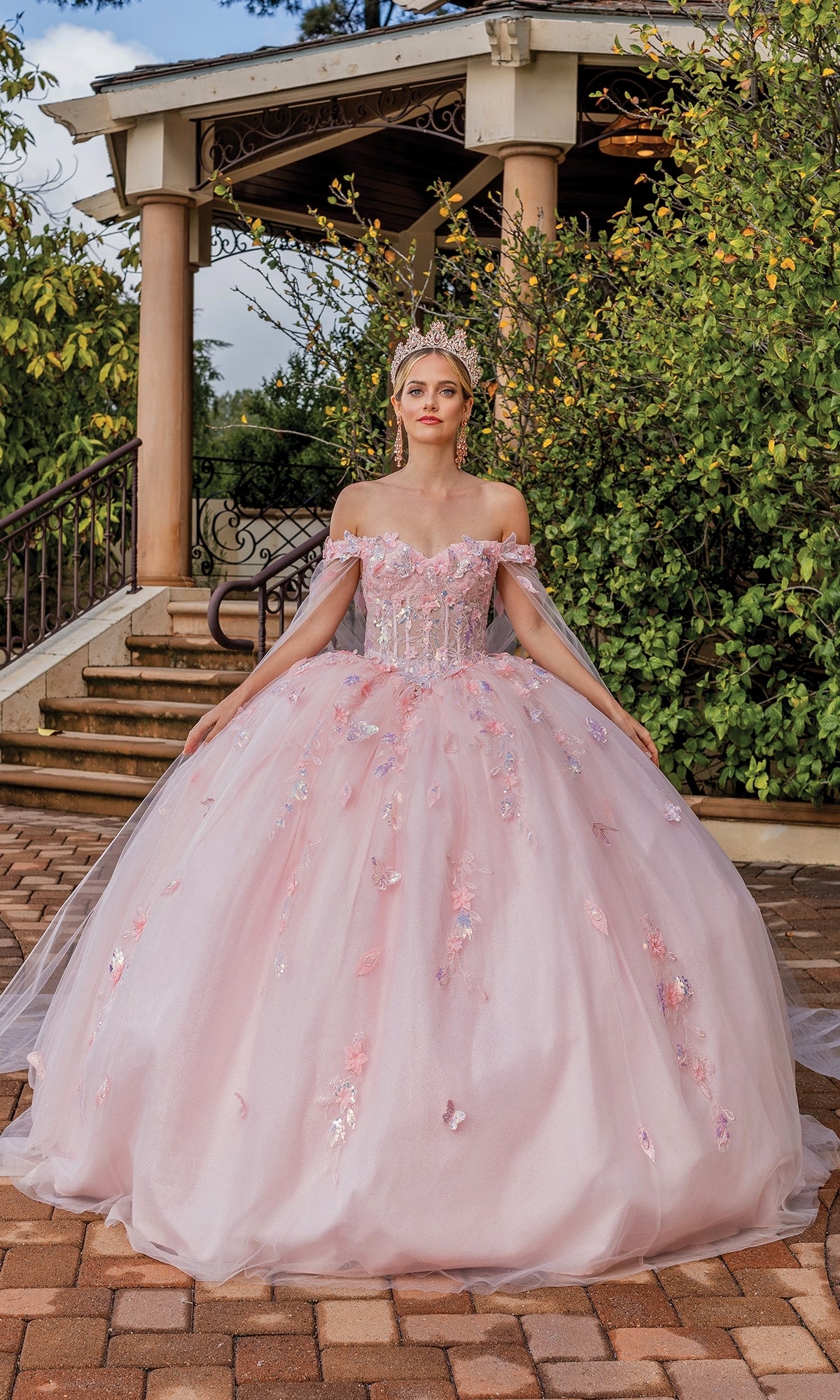 A woman in the Quinceanera Dress 1840 By Dancing Queen, featuring off-the-shoulder floral details and a tiara, stands on a brick path in front of a gazebo.